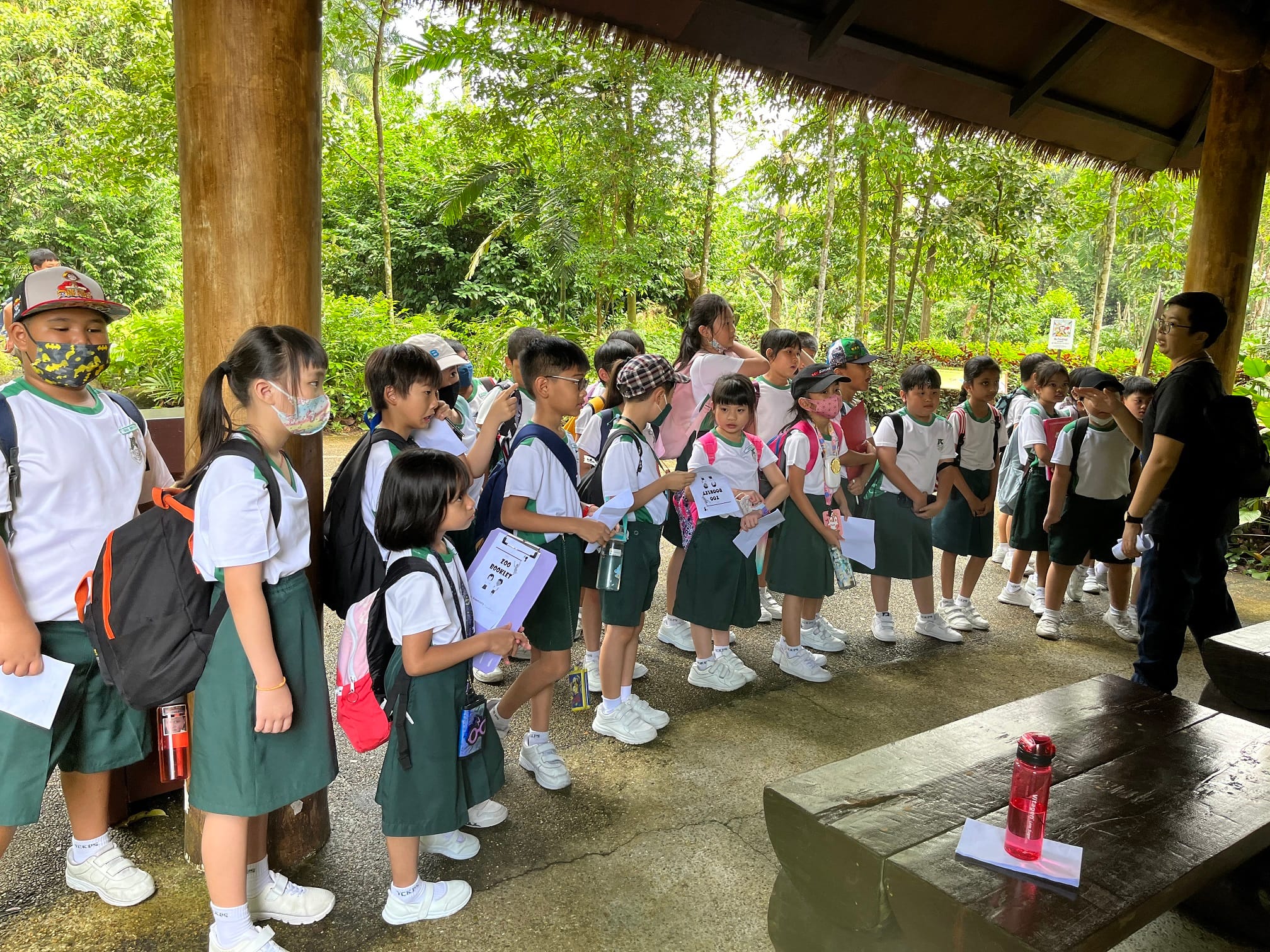 A group of young students in white and green uniforms with backpacks stand in an outdoor pavilion surrounded by trees.